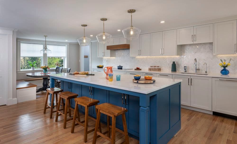 Modern kitchen with blue island, wooden stools, and elegant lighting, featuring herringbone backsplash.